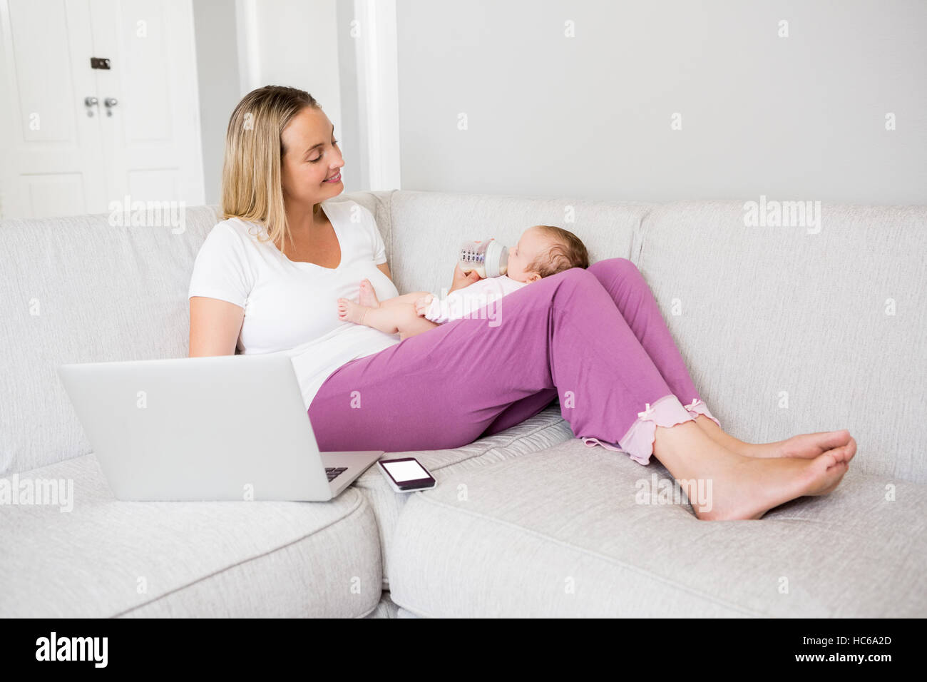 Madre utilizzando laptop mentre si alimenta il suo bambino con bottiglia di latte Foto Stock