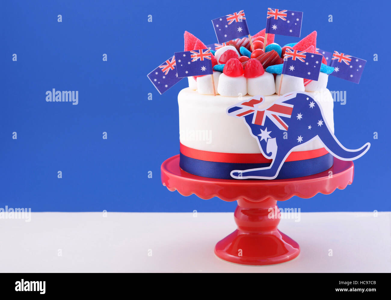 Felice l'Australia Day celebrazione torta con bandiere, marshmallow e caramelle decorazioni su una torta rosso stand su un tavolo bianco contro uno sfondo blu. Foto Stock