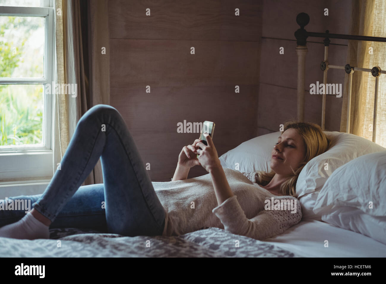 Bella donna sdraiata sul letto e utilizzando il telefono cellulare in camera da letto Foto Stock