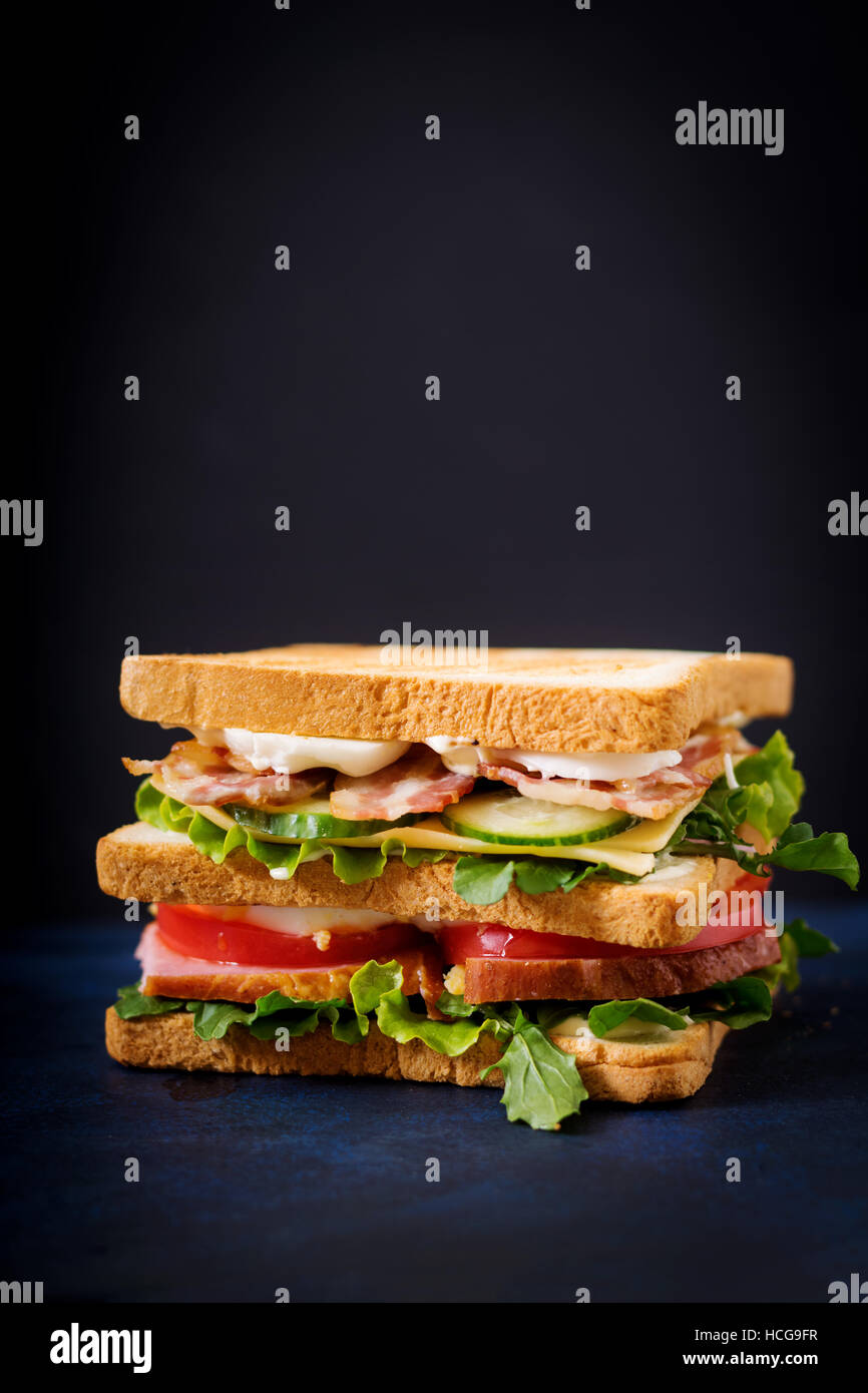 Big club sandwich al prosciutto, pancetta, pomodori, cetrioli, formaggio, uova e erbe su sfondo scuro Foto Stock