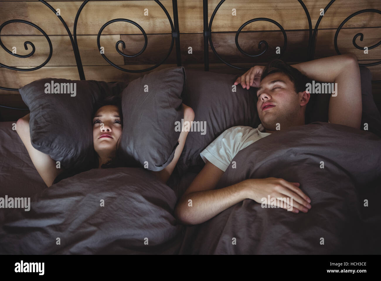 Infastiditi donna che copre le sue orecchie con cuscini per bloccare il russamento in camera da letto Foto Stock