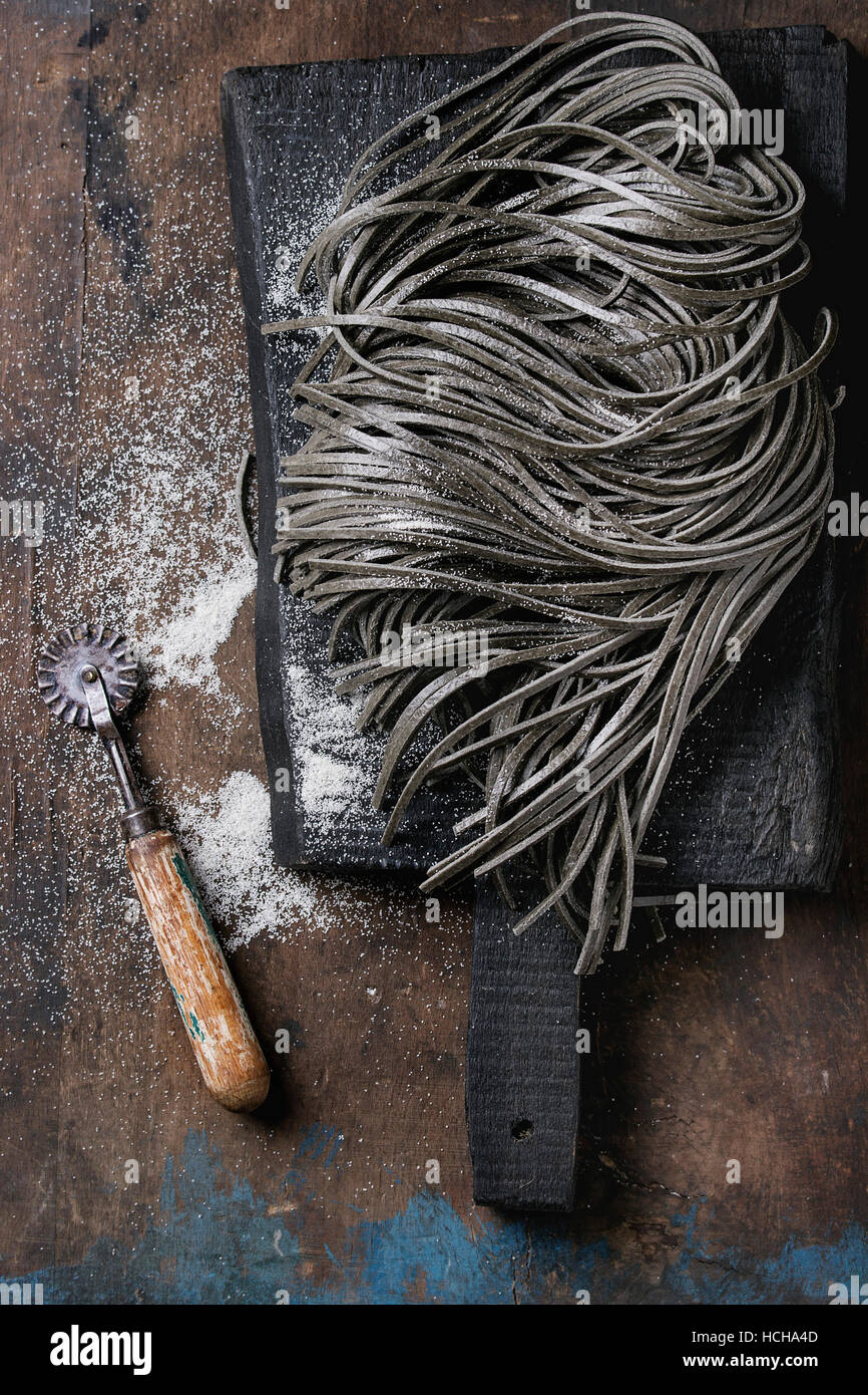 Materie non cotti nero inchiostro seppia spaghetti la pasta con la farina e il disco di pasta cutter su nero tagliere su legno scuro dello sfondo. Vista superiore con Foto Stock