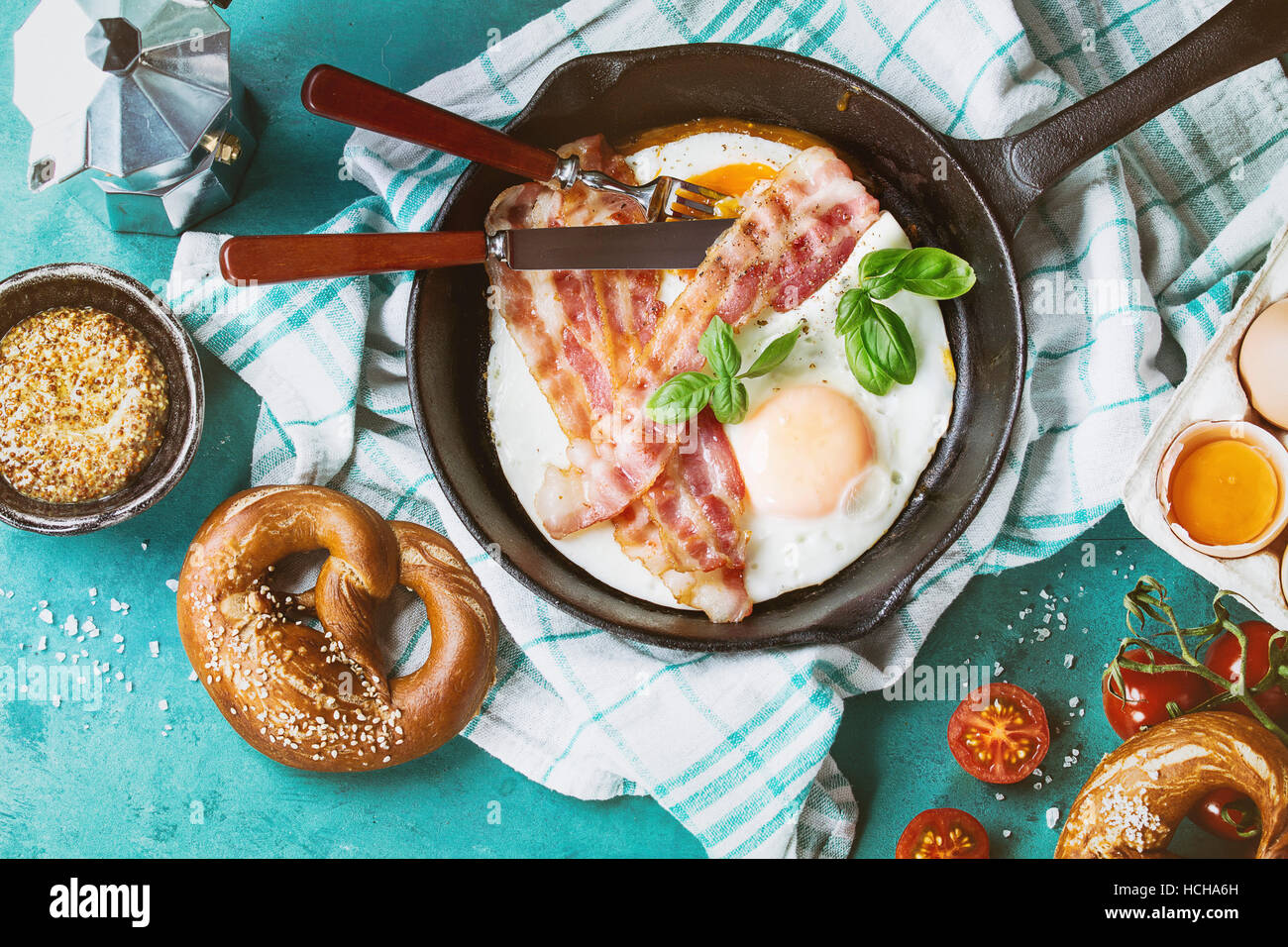 La prima colazione con uova fritte e bacon in ghisa di PAN, di uova rotte, pomodori, senape, salatini e basilico sul bianco asciugamano da cucina su legno color turchese b Foto Stock