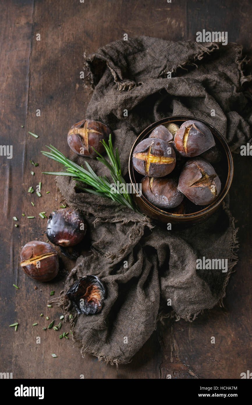 Castagne arrostite nella cenere con rosmarino in barattolo di latta su un letto di sacco su un vecchio legno scuro dello sfondo. Vista da sopra con spazio per il testo. Foto Stock