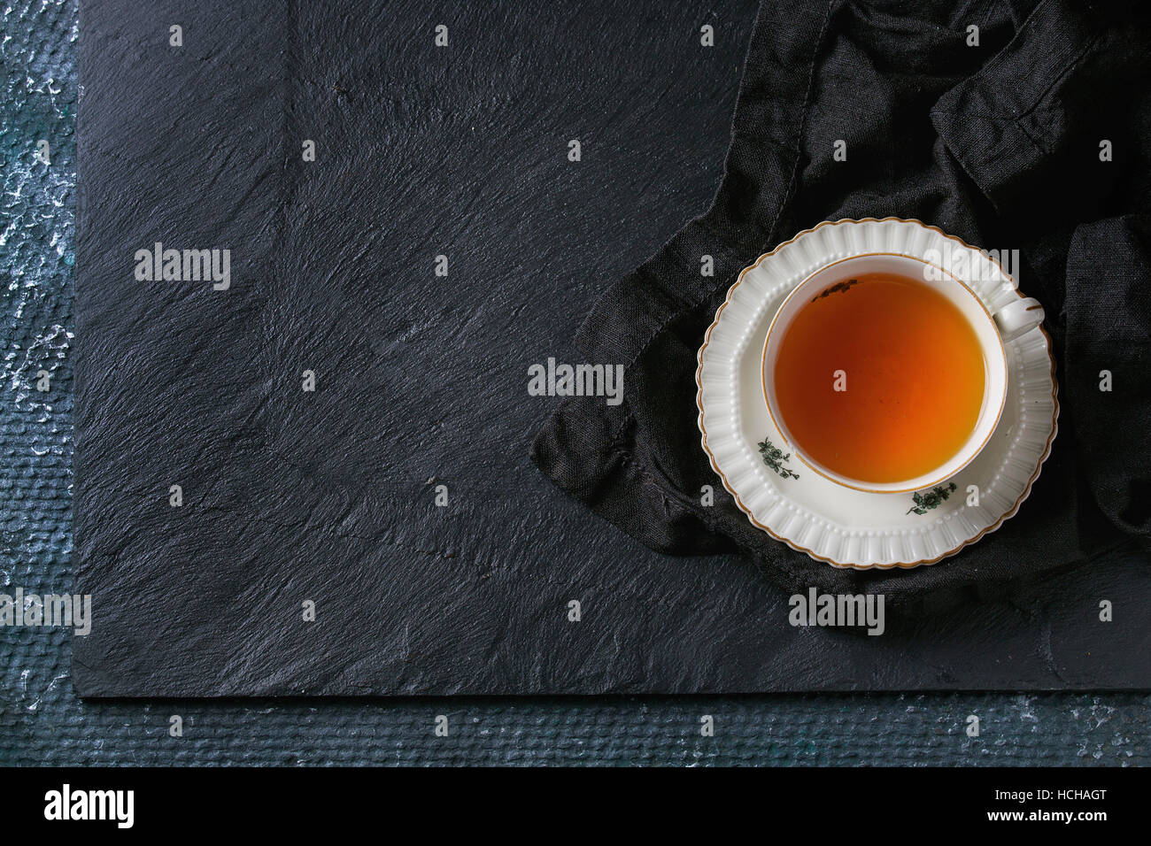Vintage bianco tazza di tè caldo sul piattino in piedi sul tessuto nero igienico sul nero ardesia sfondo texture. Vista superiore, lo spazio per il testo Foto Stock