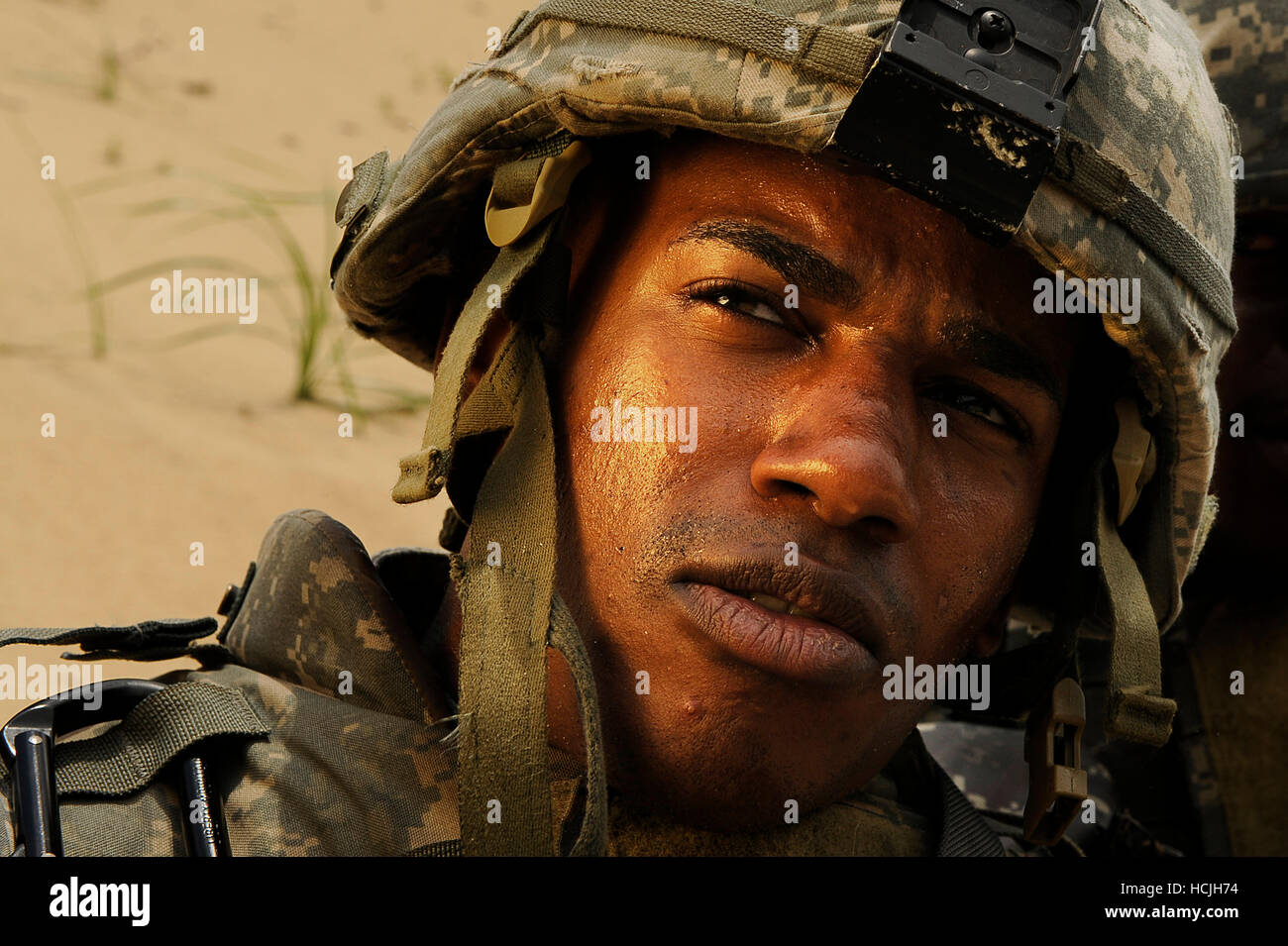 Un soldato sudato e sabbia guarda oltre la sua spalla destra. La cinghia per il mento sul suo casco di mimetizzazione è sganciato e il collare della sua armatura per il corpo è coperto di sporcizia. Foto Stock