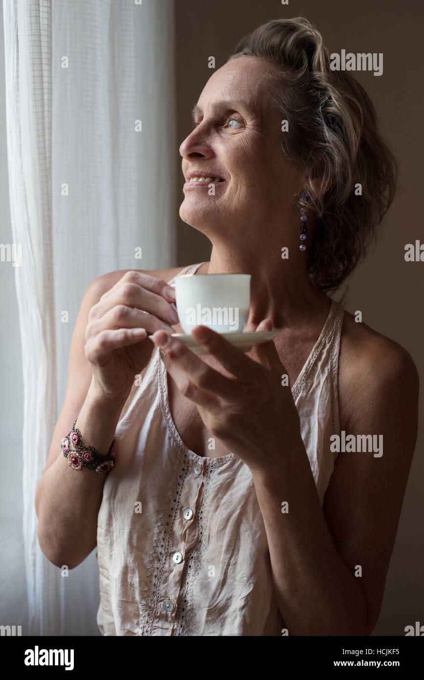Donna matura in piedi presso la finestra con una tazza di tè Foto Stock