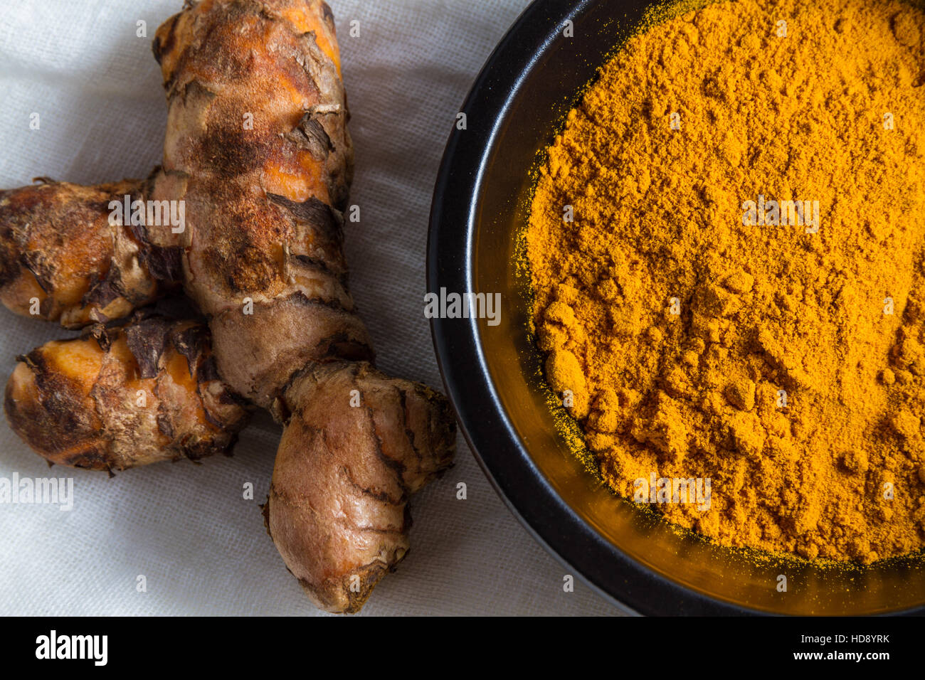 Pezzo di curcuma radice o Curcuma longa dalla ciotola di curcuma in polvere sul panno bianco sfondo, macro. Foto Stock