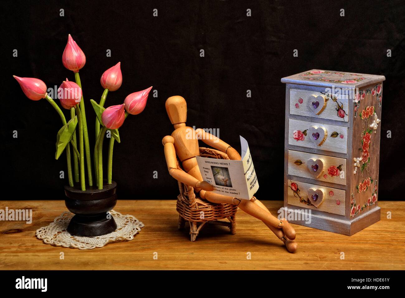 Figura in legno leggendo il giornale in soggiorno, accanto a cassettiera e fiori in legno con sfondo scuro Foto Stock