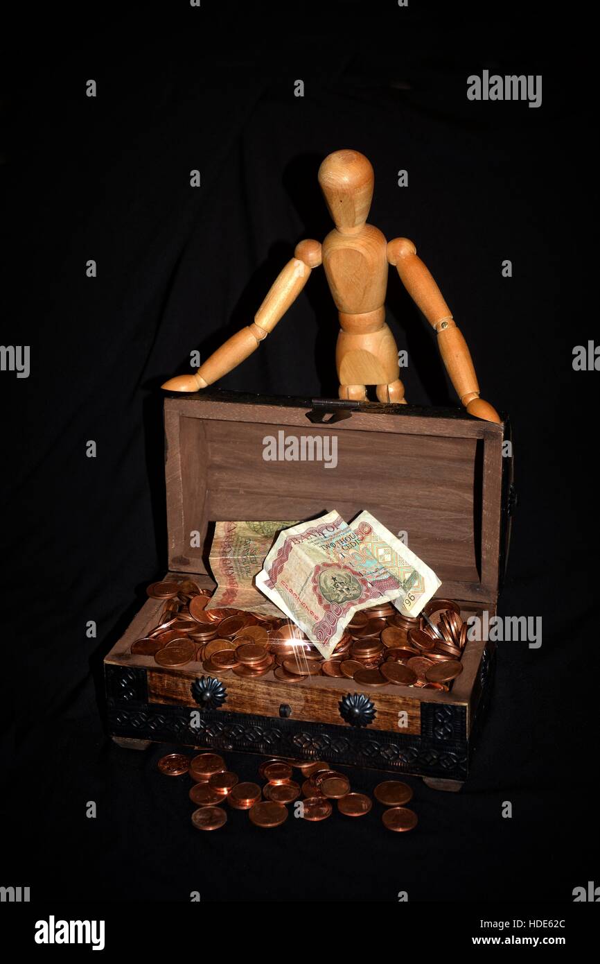 La figura di legno in piedi dietro una scatola di legno piena di soldi - monete contro lo sfondo nero Foto Stock