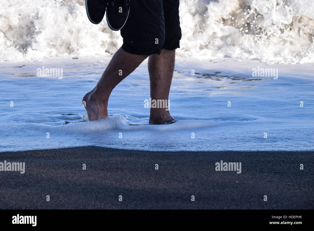 Messa a fuoco su un uomo che trasportava formatori passeggiando attraverso il surf in bordo di mare Foto Stock