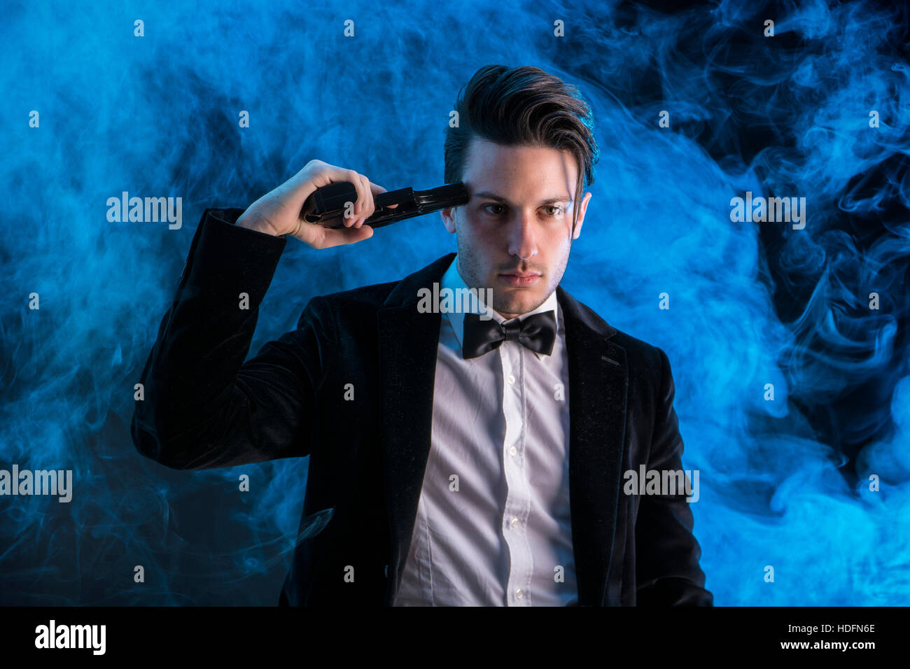 Giovane uomo bello in tuta tenendo la pistola al tempio e guardando dritto su sfondo nero con il fumo. Foto Stock