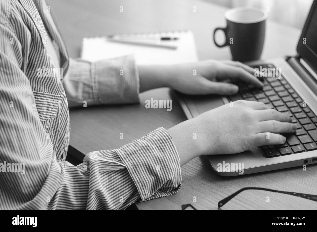 Mani femminili lavorando a un computer portatile, immagine in bianco e nero. Tazza da caffè, notebook, penna, matita, occhiali Foto Stock