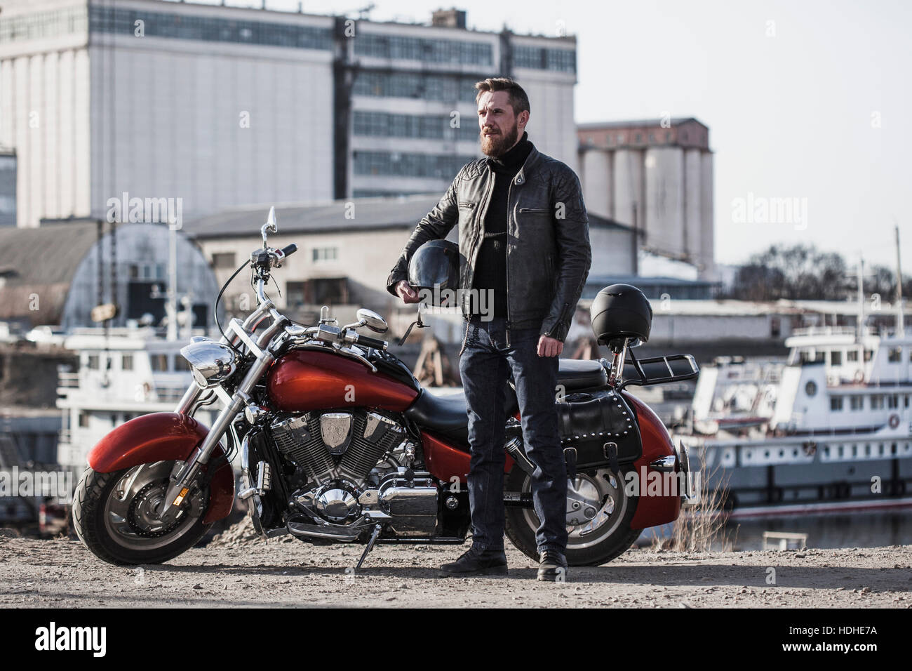 A piena lunghezza Ritratto di biker holding casco mentre in piedi da moto contro l'impostazione industriale Foto Stock