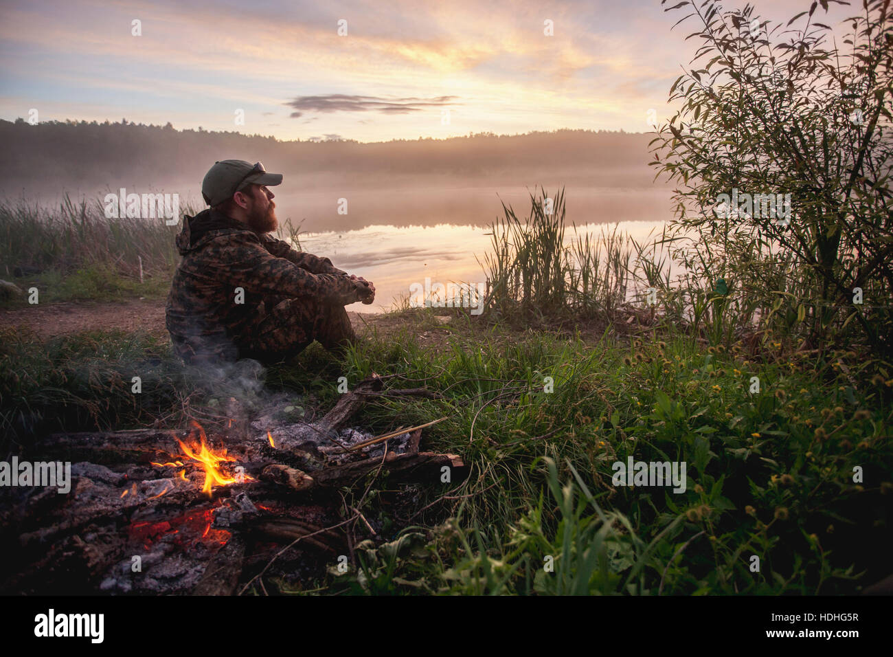 Vista laterale del cacciatore seduto da falò in campo al lungolago durante il tramonto Foto Stock
