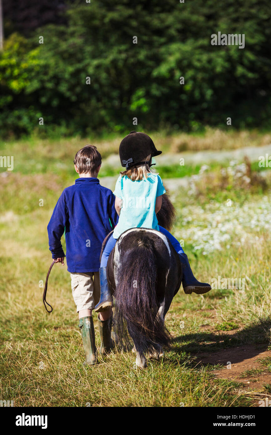 Una ragazza in sella ad un pony e un ragazzo a piedi attraverso un campo. Foto Stock
