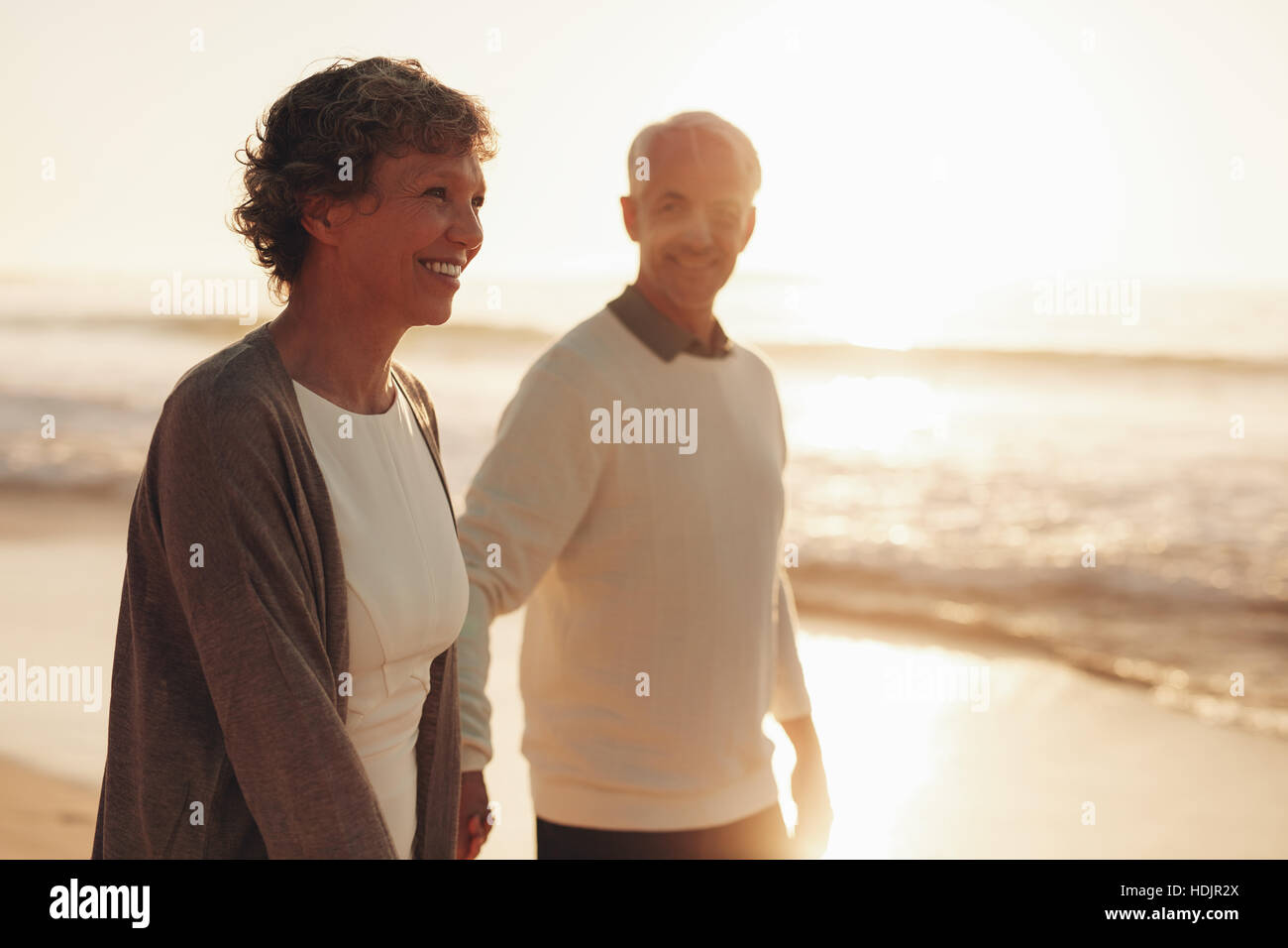 Senior sorridente Donna che cammina con il suo marito sulla spiaggia al tramonto. Coppia matura camminando lungo la riva del mare. Foto Stock