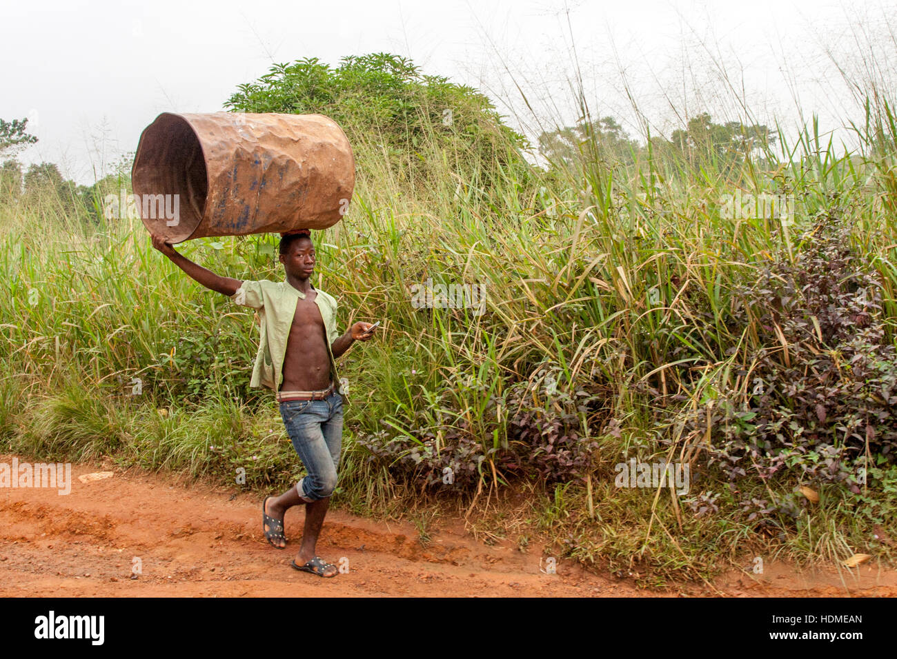 Multitasking in Sierra Leone: Uomo che porta un Barrel sulla testa mentre usa il suo smartphone. Il barile di petrolio ha fatto molta strada in Africa ed è ancora prezioso nonostante le sue ammaccature Foto Stock