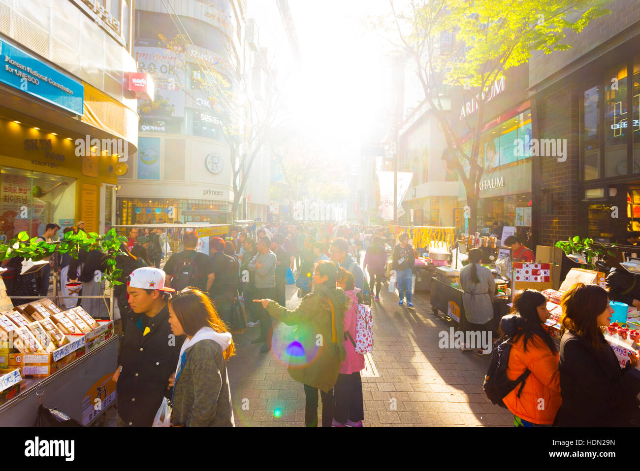 Sun le retroilluminazioni turisti camminando giù a Myeongdong trafficata strada pedonale dello shopping con commercialismo di negozi, segni e affollato Foto Stock