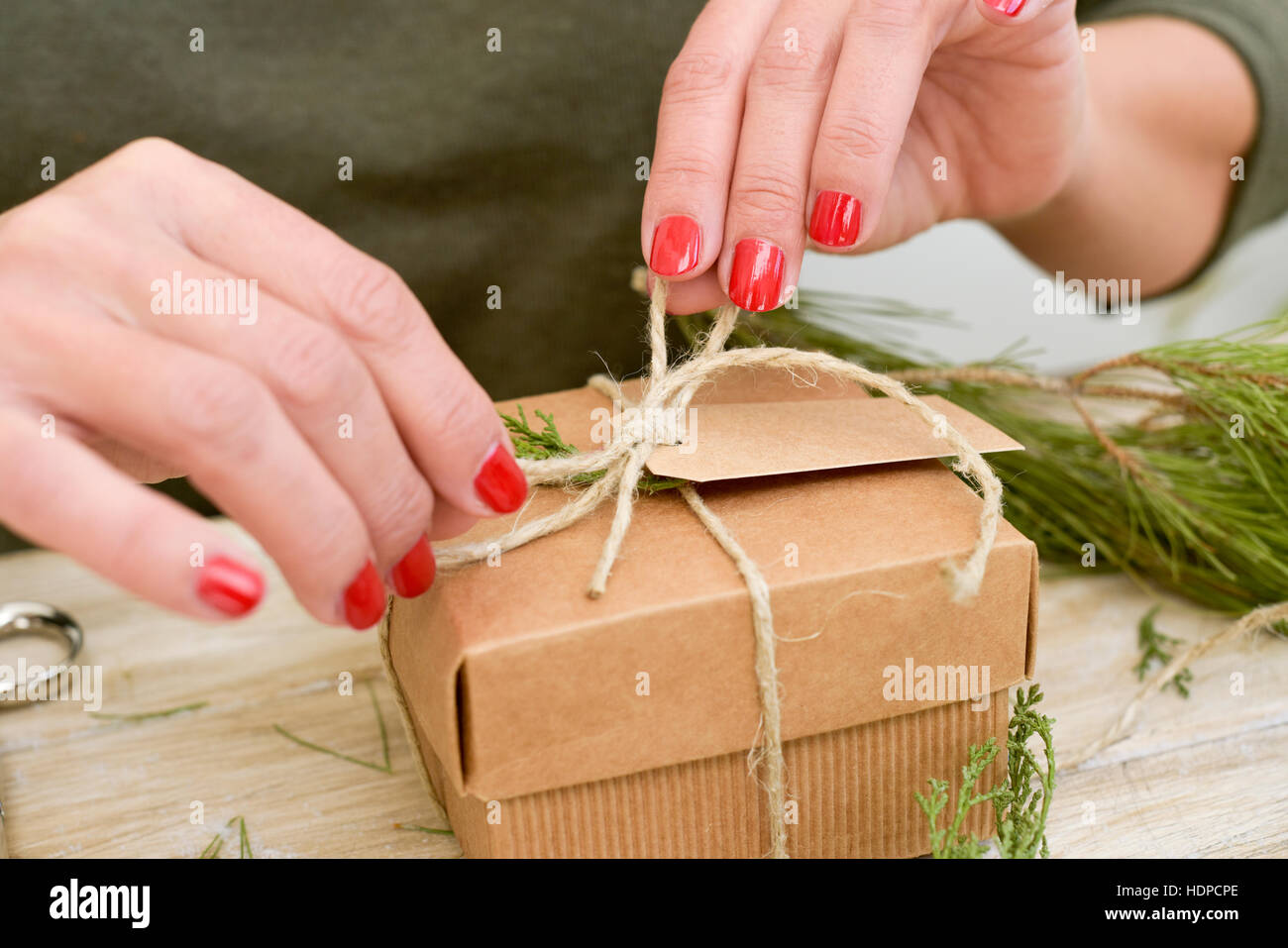 Primo piano di una giovane donna caucasica con lei le unghie dipinte di rosso legatura di una corda di iuta attorno ad un cartone marrone confezione regalo su un rustico di una superficie di legno Foto Stock