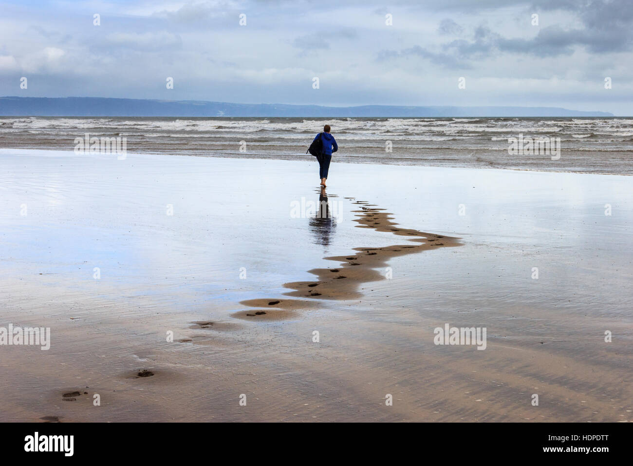 Una donna camminare a piedi scalzi verso il mare su una spiaggia di spazzate dal vento lasciando tracce nella sabbia, Condino, Devon, Inghilterra, Regno Unito Foto Stock