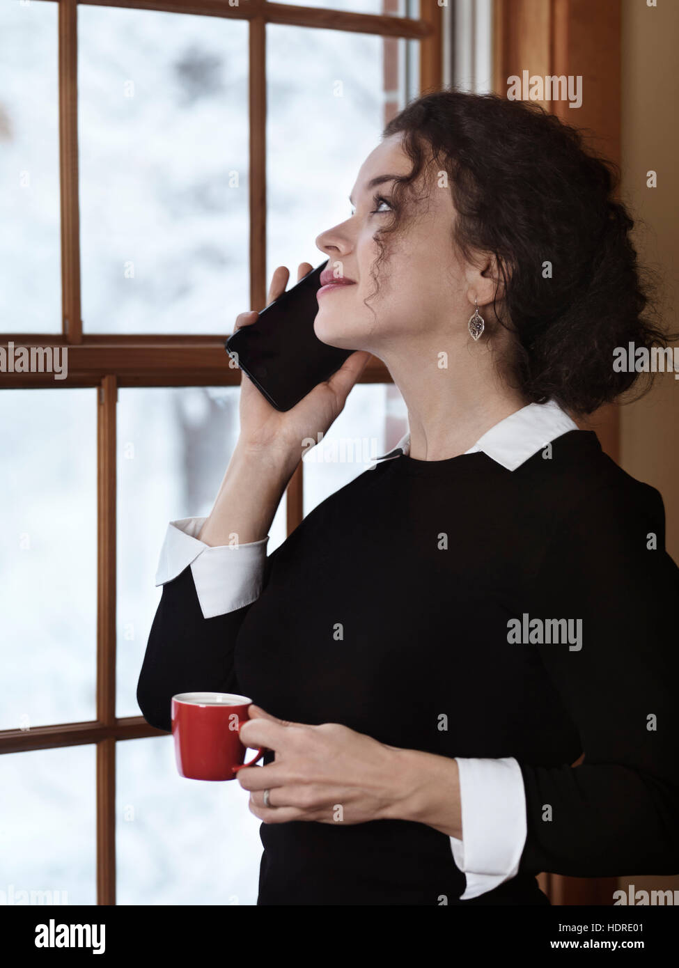 Donna con una tazza di caffè a parlare al telefono da una finestra con un innevato paesaggio invernale dietro di esso. Apple iPhone 7 plus. Foto Stock