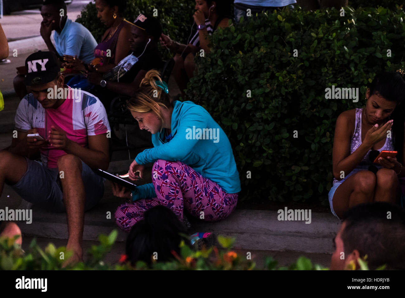 Le persone sui loro telefoni cellulari e compresse in corrispondenza di una zona wifi in un parco al tramonto a Cienfuegos, Cuba Foto Stock