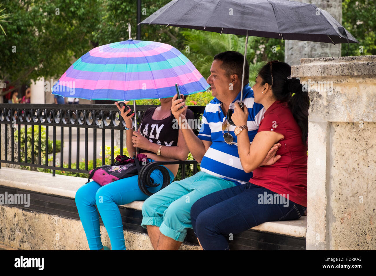 Le persone sotto gli ombrelloni a wifi zone sui loro telefoni cellulari in una plaza a Camaguey, Cuba Foto Stock
