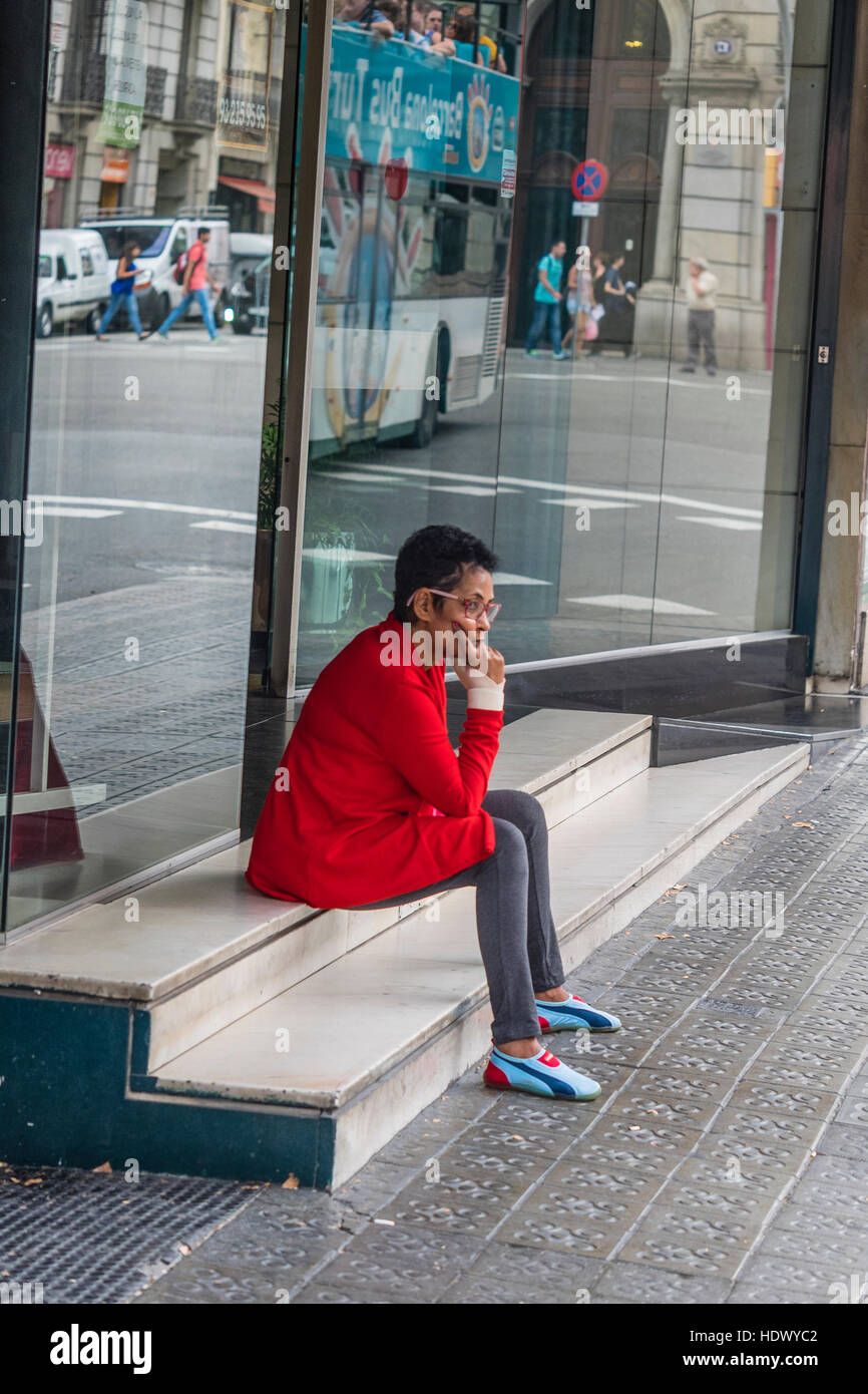 Una femmina spagnolo vestito con una giacca rossa e voglia di scarpe da corsa e collant, si siede sui gradini di marmo al di fuori di un negozio di fronte in attesa in Barcellona, Spagna. Foto Stock