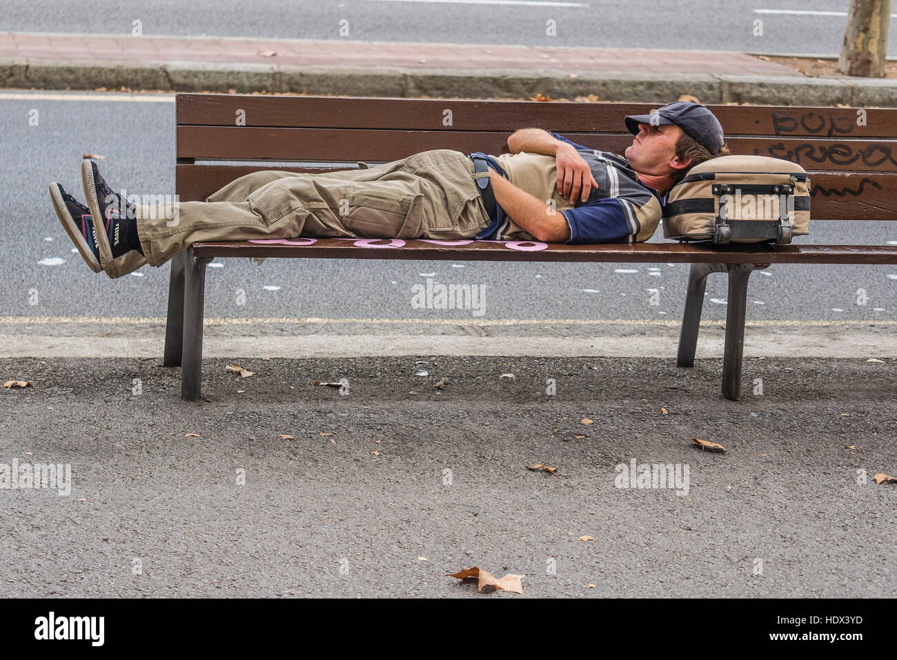 Napping maschio su un banco della città di Barcellona, in Spagna con il suo cappello sul suo volto e la sua testa in appoggio sul suo zaino. Foto Stock