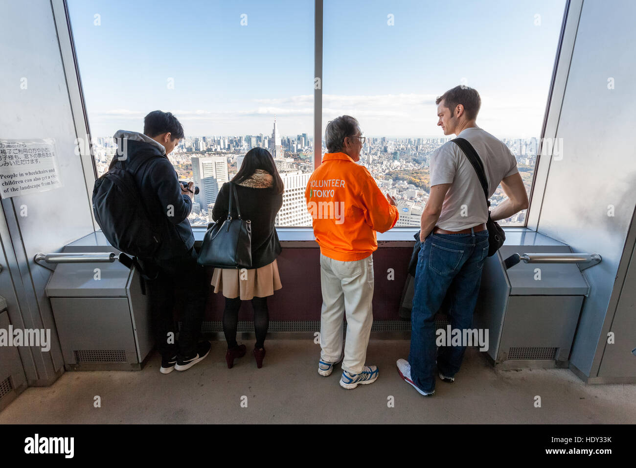 Una guida turistica volontaria (in giacca arancione) guida un turista maschile bianco intorno al ponte di osservazione al 45th° piano della torre TMG. Shinjuku, Tokyo, Giappone Foto Stock