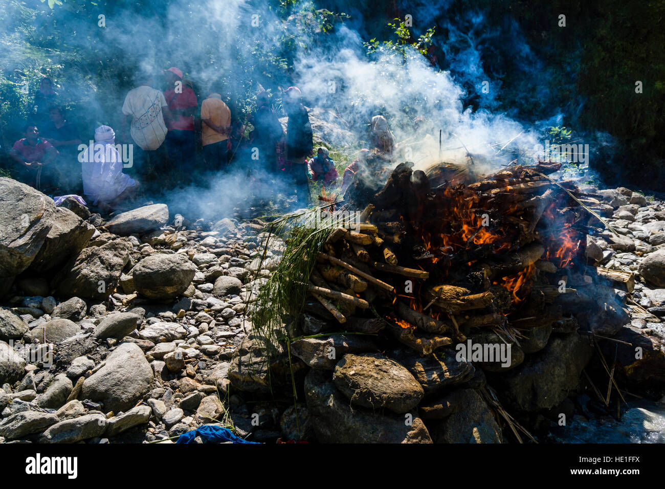 Il fuoco del funerale di un corpo di morte al suolo di cremazione sulla banca del fiume Kali Gandaki è produrre un sacco di smok, le persone sono seduta b Foto Stock