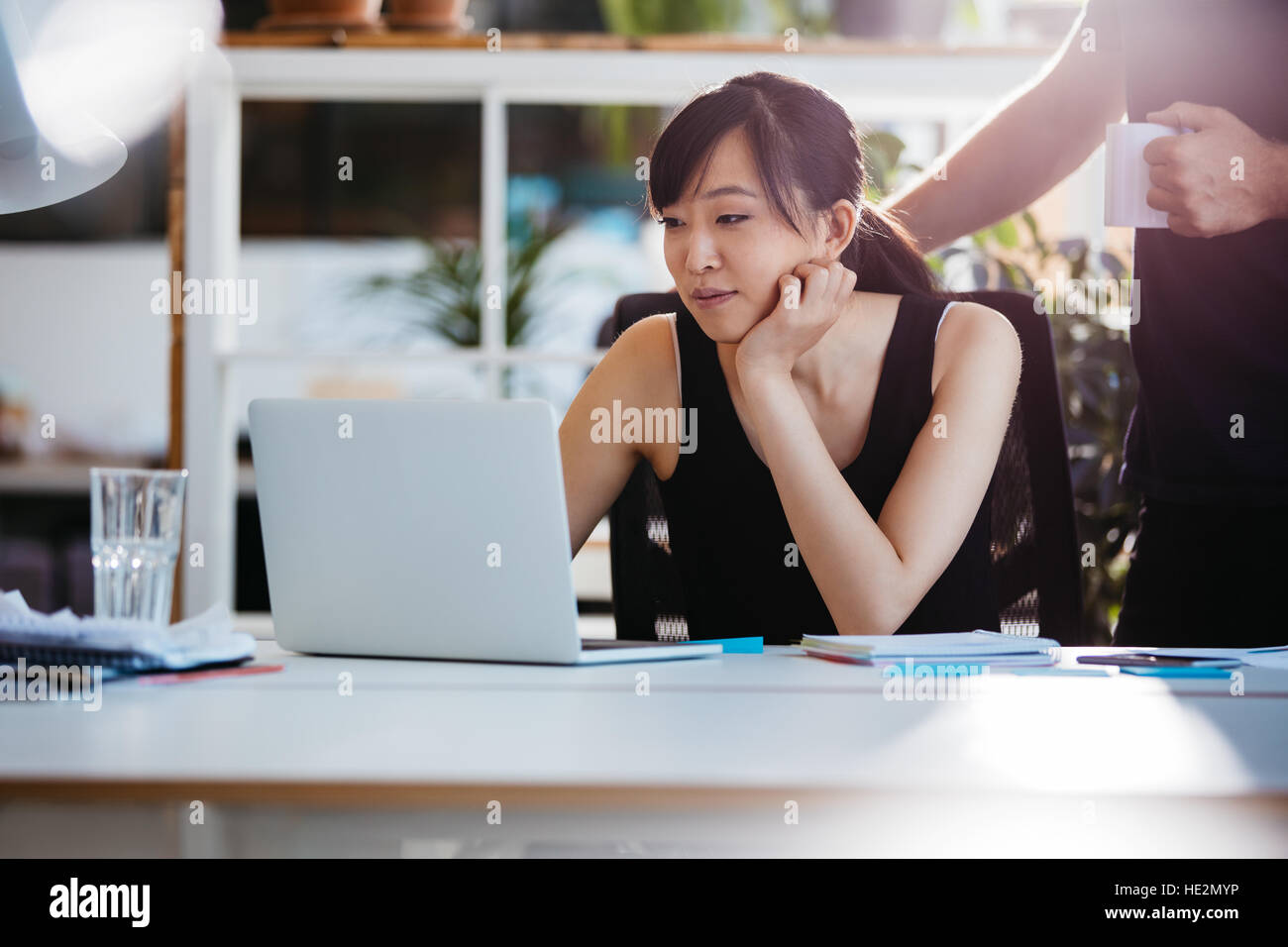 Colpo di asian giovane donna seduta alla sua scrivania che lavora sul computer portatile con la collega di sesso maschile in attesa. Foto Stock
