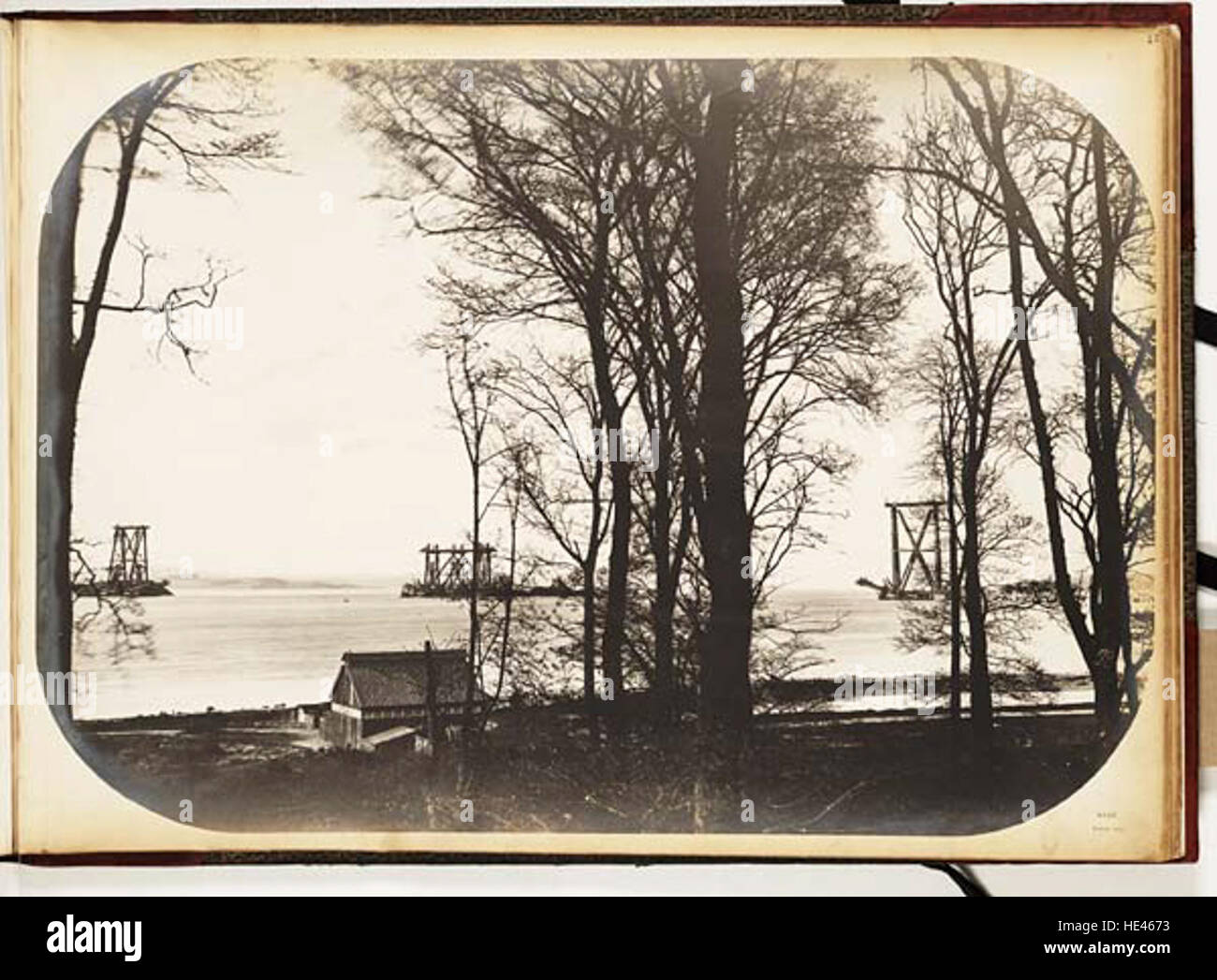 Questa fotografia storica mostra la costruzione del Forth Bridge da Port Edgar, catturando la monumentale impresa ingegneristica. L'immagine evidenzia gli alberi circostanti, aggiungendo un contesto al paesaggio dell'epoca. La foto riflette sia il progresso industriale che l'ambiente naturale del tempo. Foto Stock