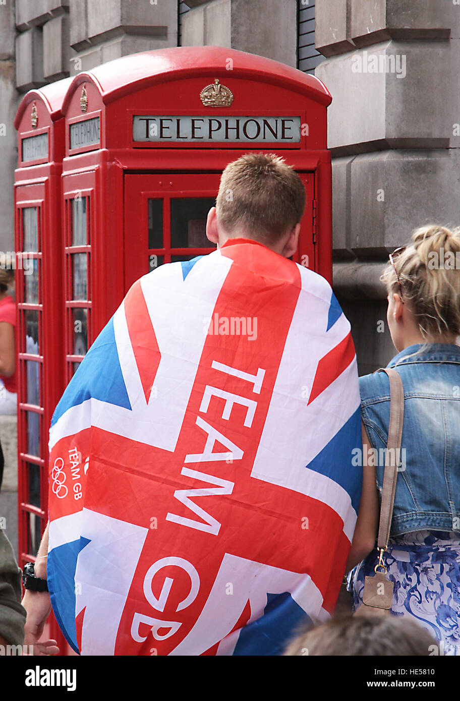 Appassionati di sport con unione jack intorno alle sue spalle con il team GB stampati su di essa a piedi passato telefono rosso chiosco nel 2012 Foto Stock
