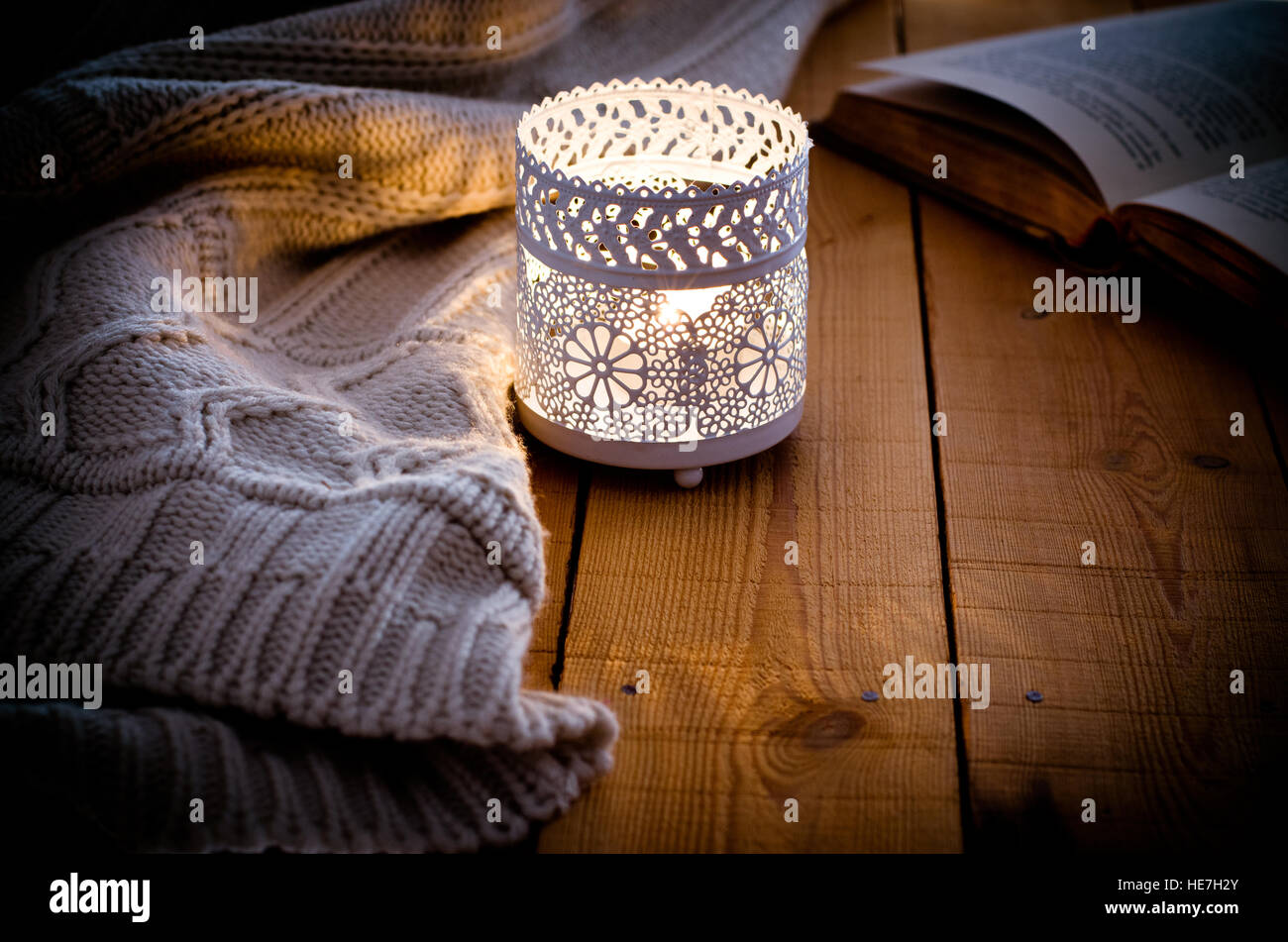 Candela accesa in un merletto portacandele, felpa lavorata a maglia e libro aperto su uno sfondo di legno, accogliente winter o serata autunnale Foto Stock