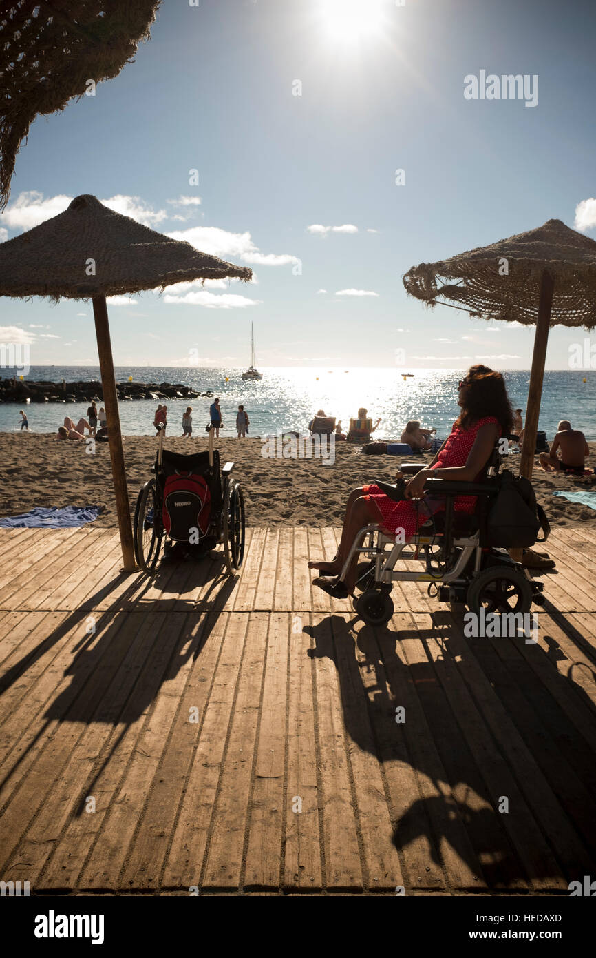 La donna in una sedia a rotelle di alimentazione fotografato su un ponte di legno sul lato ammirate una spiaggia soleggiata con la gente a prendere il sole Foto Stock