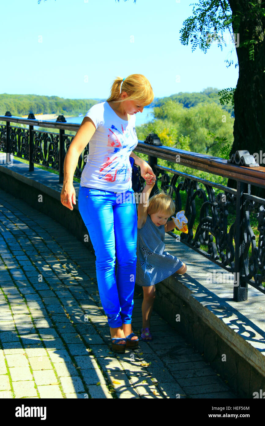 Moderna moda donna conduce la sua piccola figlia per mano sul cordolo in un parco della città Foto Stock