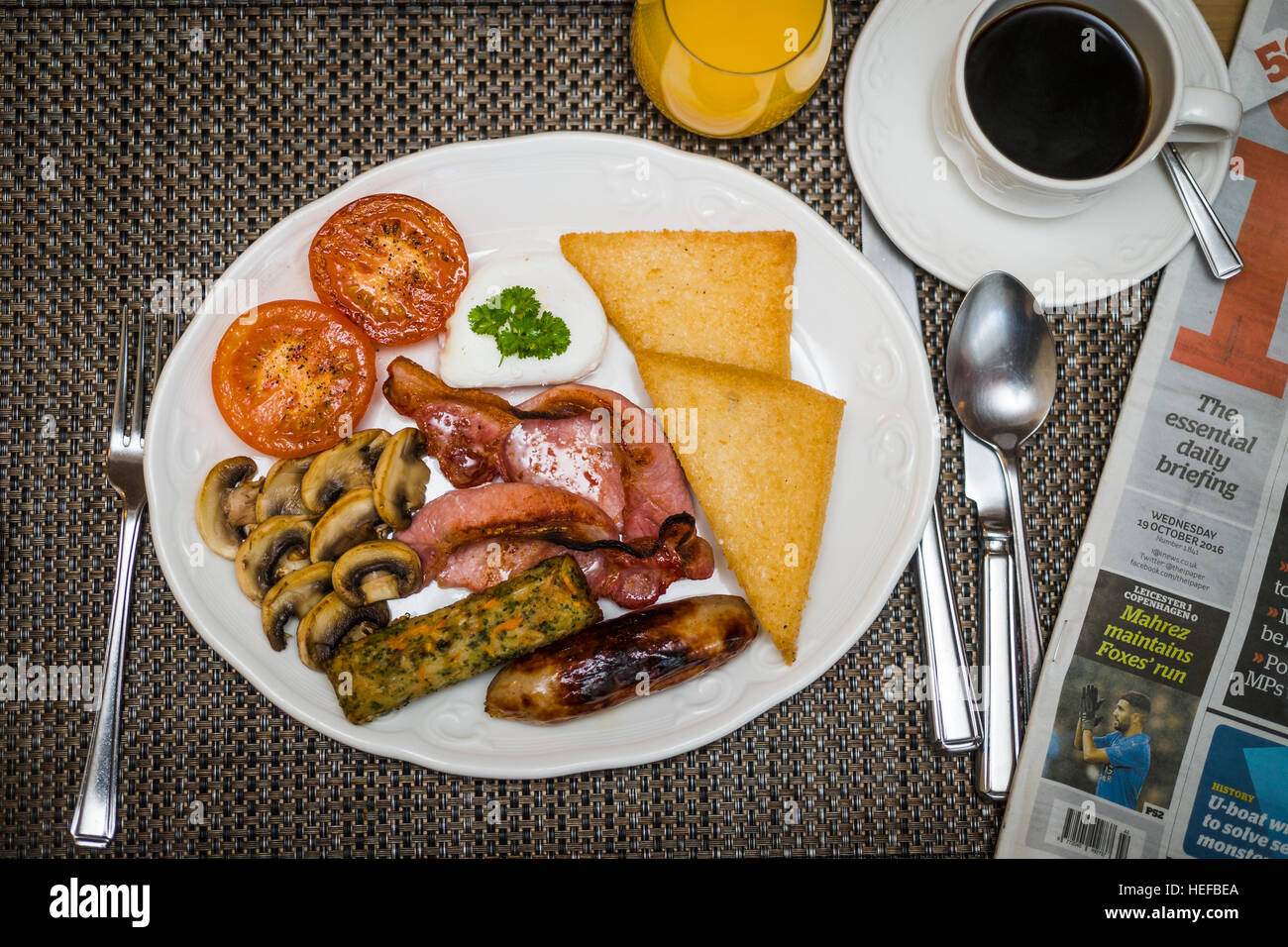 Full Monty: un fritto colazione inglese completa su una piastra bianca in un hotel del Regno Unito (pancetta, uova, salsicce, pomodori e funghi, pane fritto,) Tazza di caffè, succo d'arancia e un newpaper Foto Stock