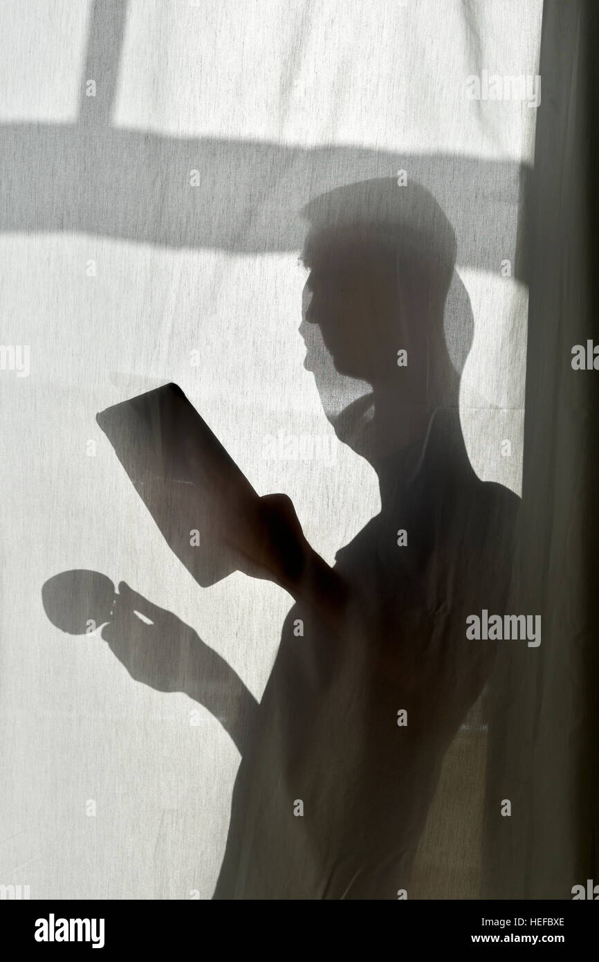 La silhouette di un giovane uomo dietro una tenda bianca con una tazza di caffè o di tè in una mano e una compressa nell'altra mano Foto Stock