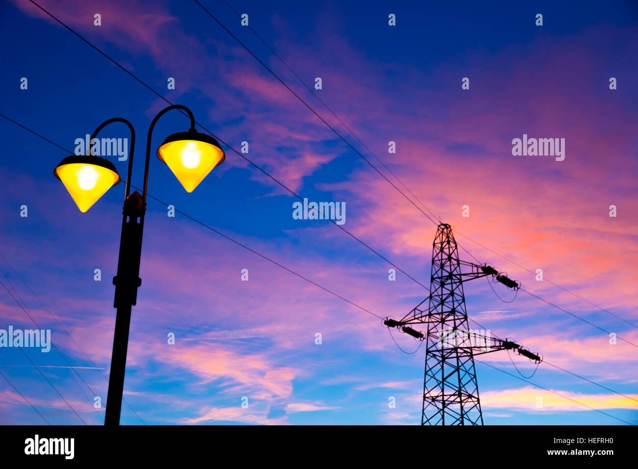 Accesa sul lampione al tramonto con linea ad alta tensione e il pilone sullo sfondo Foto Stock