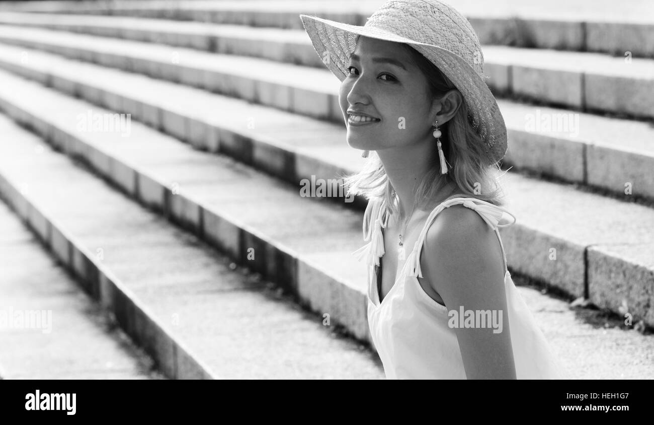 Felice, bella ragazza giapponese che indossa un cappello di paglia e sorridente è seduta sulle scale in un parco Foto Stock