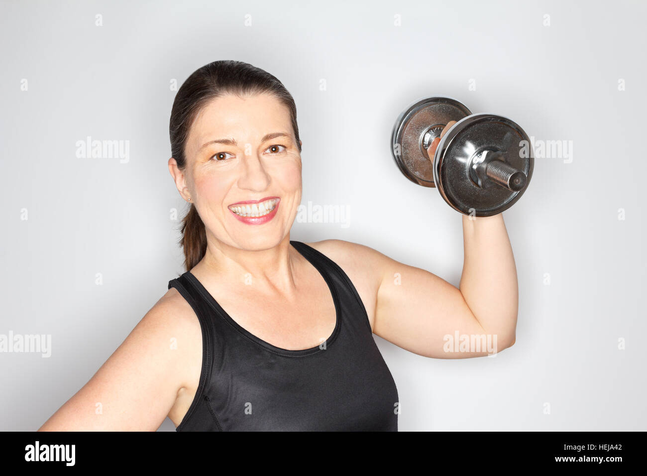 Donna di mezza età in un nero sport top il sollevamento di un carico pesante dumbbell, sorridendo felice e orgoglioso, sfondo luminoso, spazio di copia Foto Stock