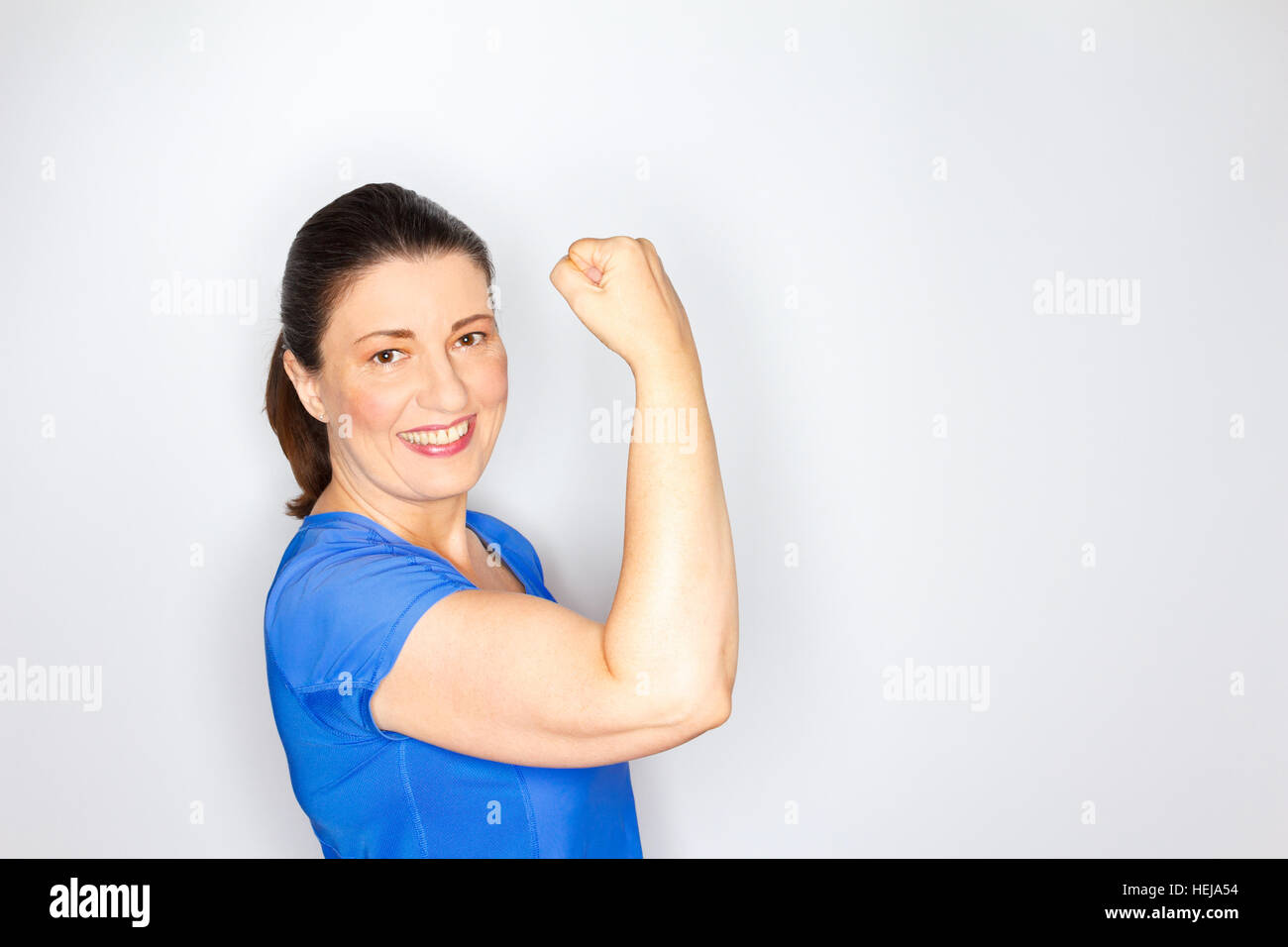 Donna di mezza età in blu luminoso abbigliamento sportivo che mostra i suoi muscoli bisceps, sfondo luminoso, copia o lo spazio di testo, copyspace Foto Stock