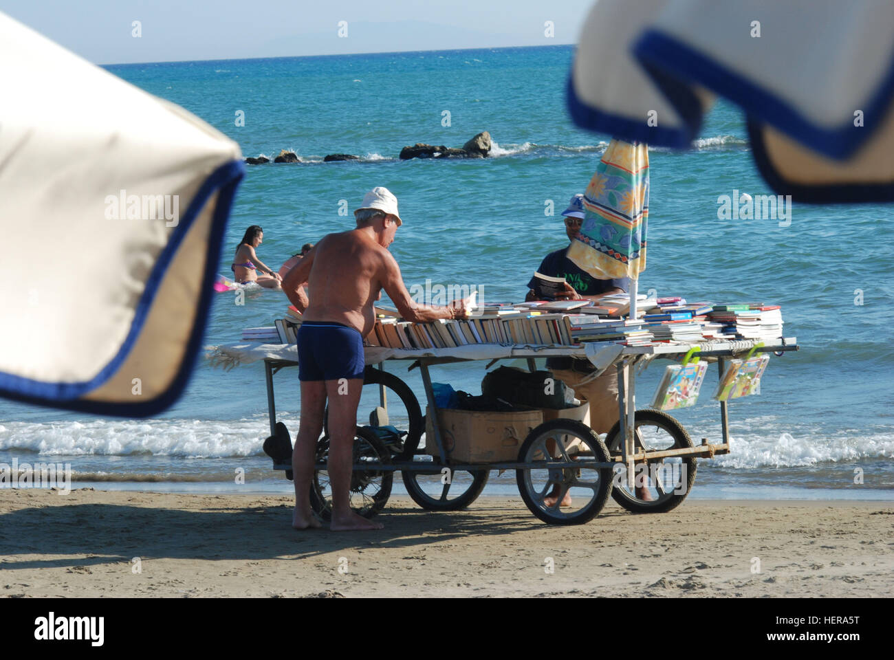 Ã"lterer Herr mit Badehose und Strandhut betrachtet ein Buch von einem fahrenden Buchladen mit gebrauchten BÃ¼chern am Strand von Castiglione della pe Foto Stock