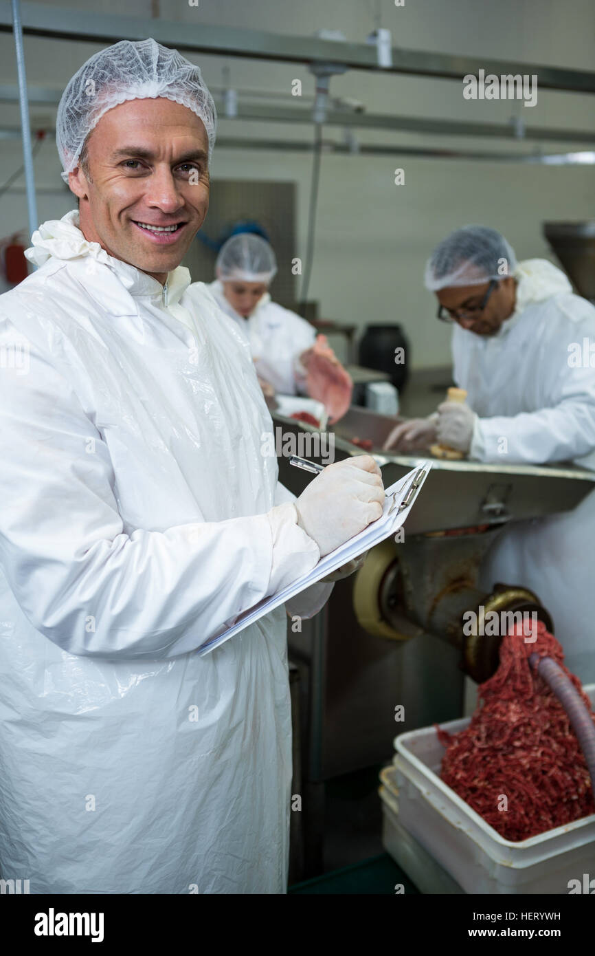 Butcher iscritto sulla clipboard mentre i colleghi ponendo la carne nel tritacarne in fabbrica a base di carne Foto Stock