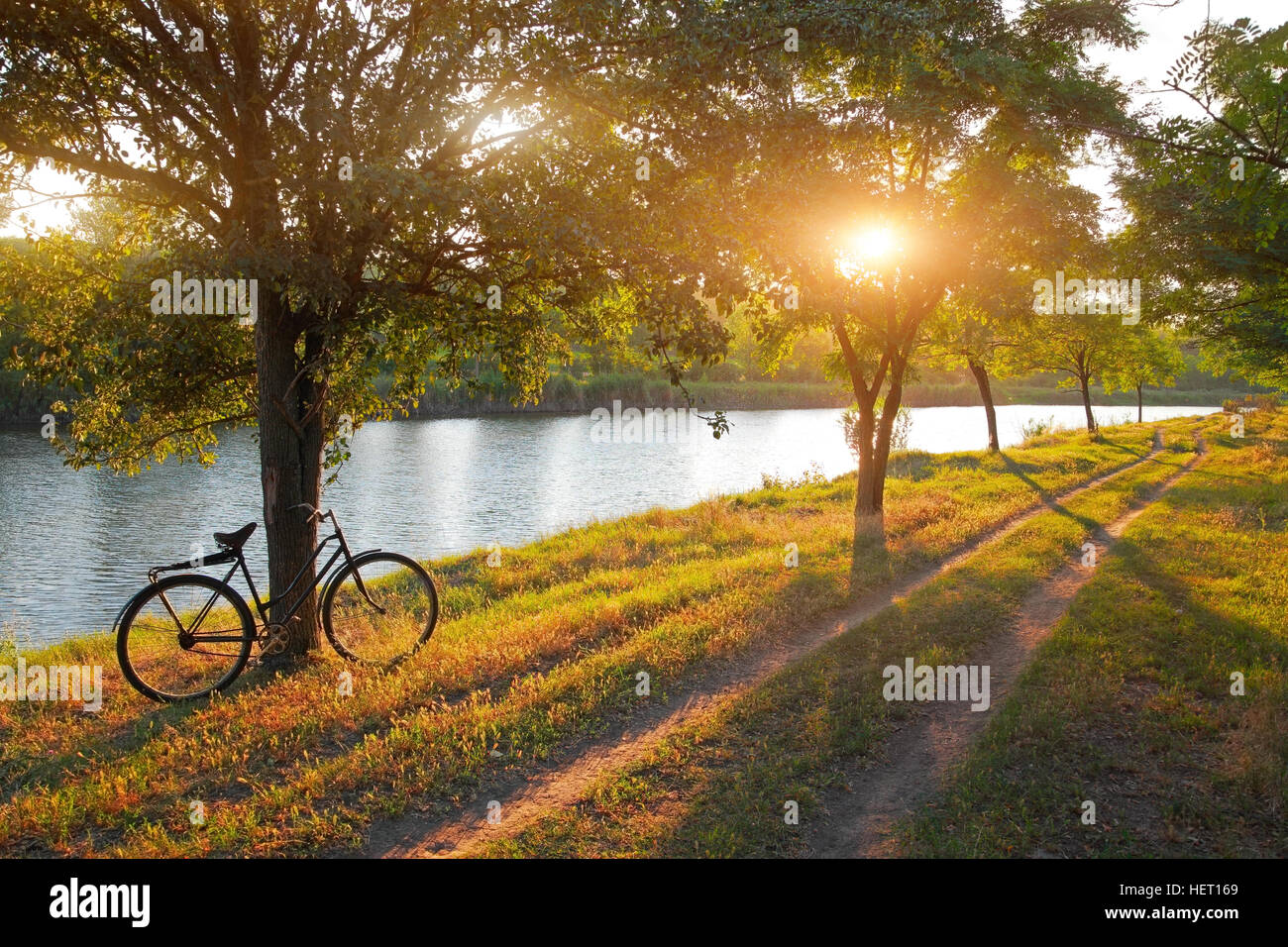 Paesaggio estivo con la bicicletta, strada rurale lungo il fiume tra gli alberi al tramonto Foto Stock