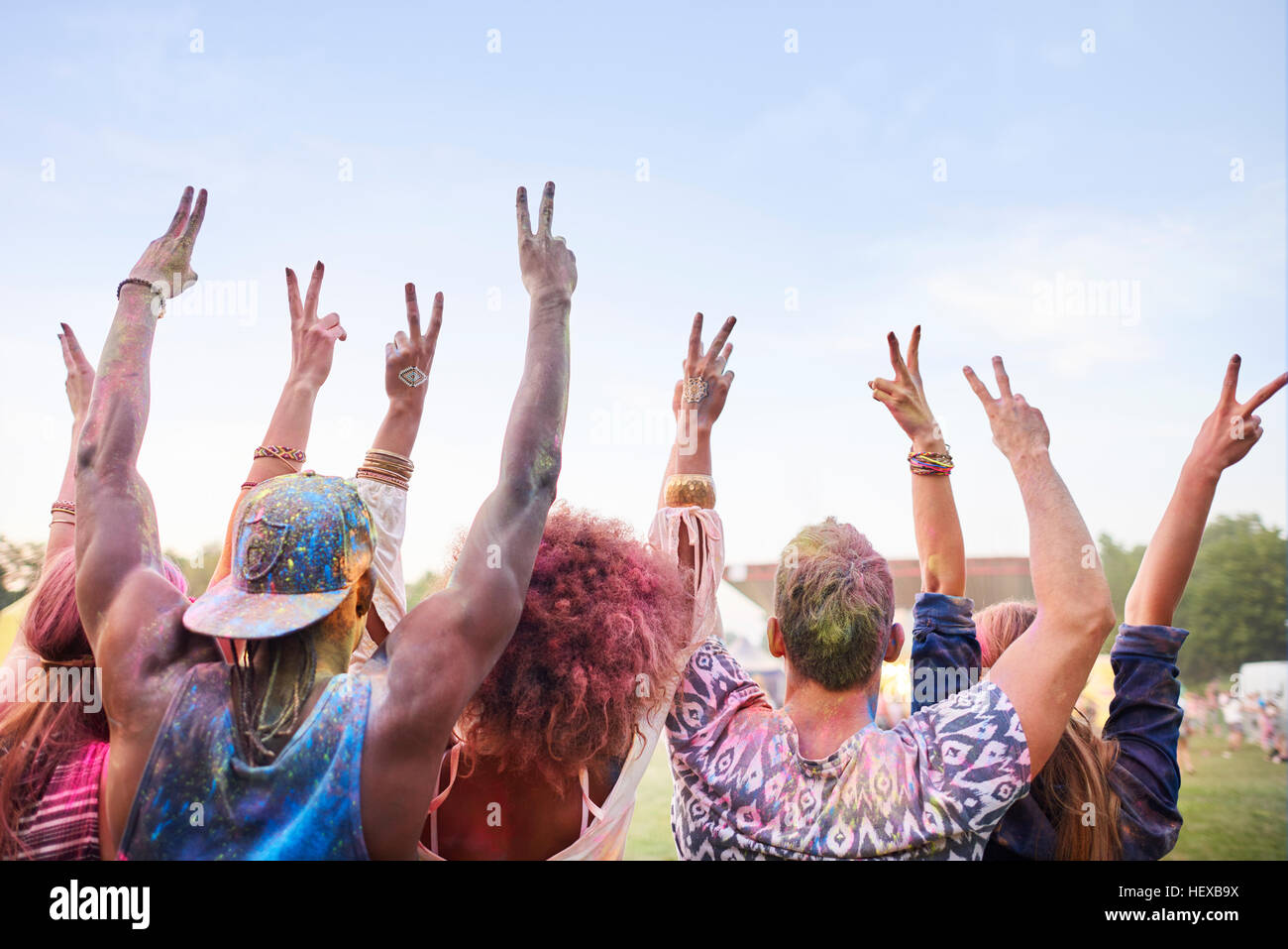 Gruppo di amici al festival, coperto di polvere colorata vernice, bracci sollevati, facendo segno di pace con le dita, vista posteriore Foto Stock