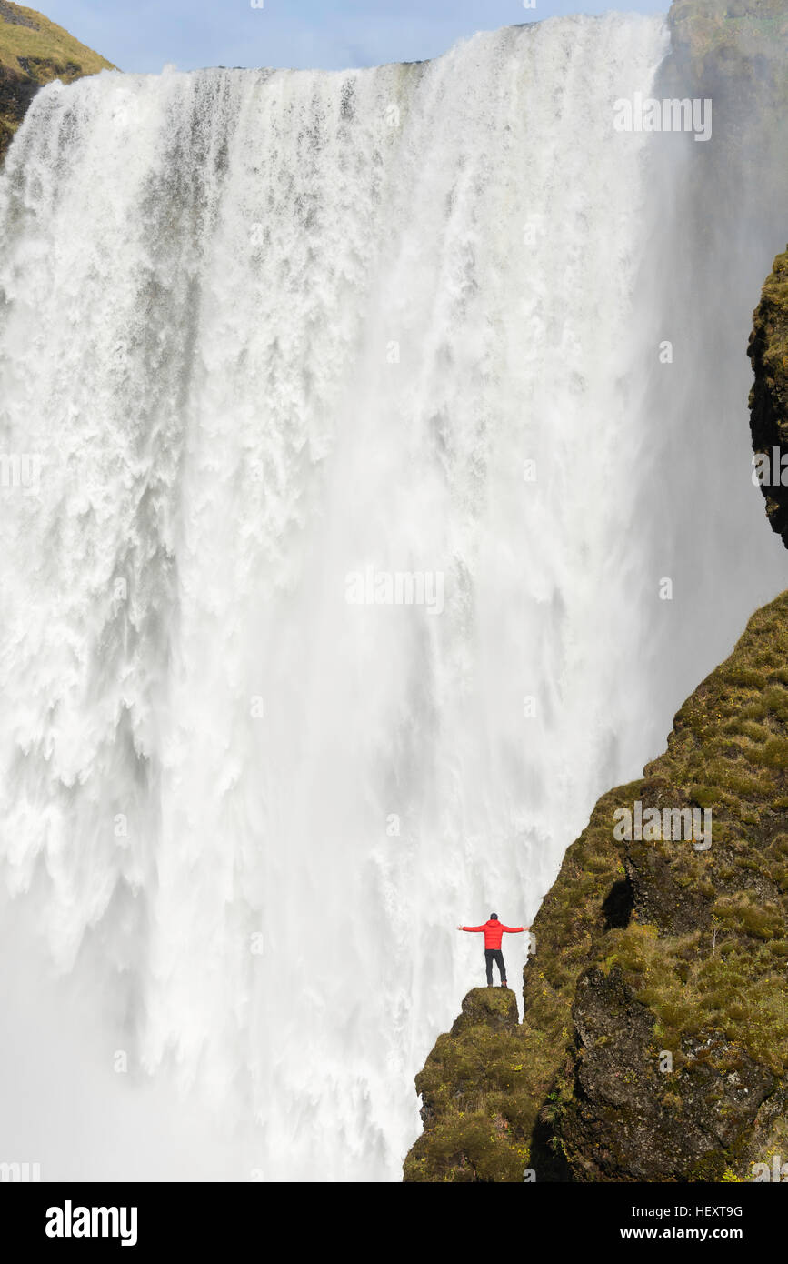 Giovane Uomo in camicia rossa sorge su una scogliera con Skogafoss cascata dietro, Islanda. Foto Stock