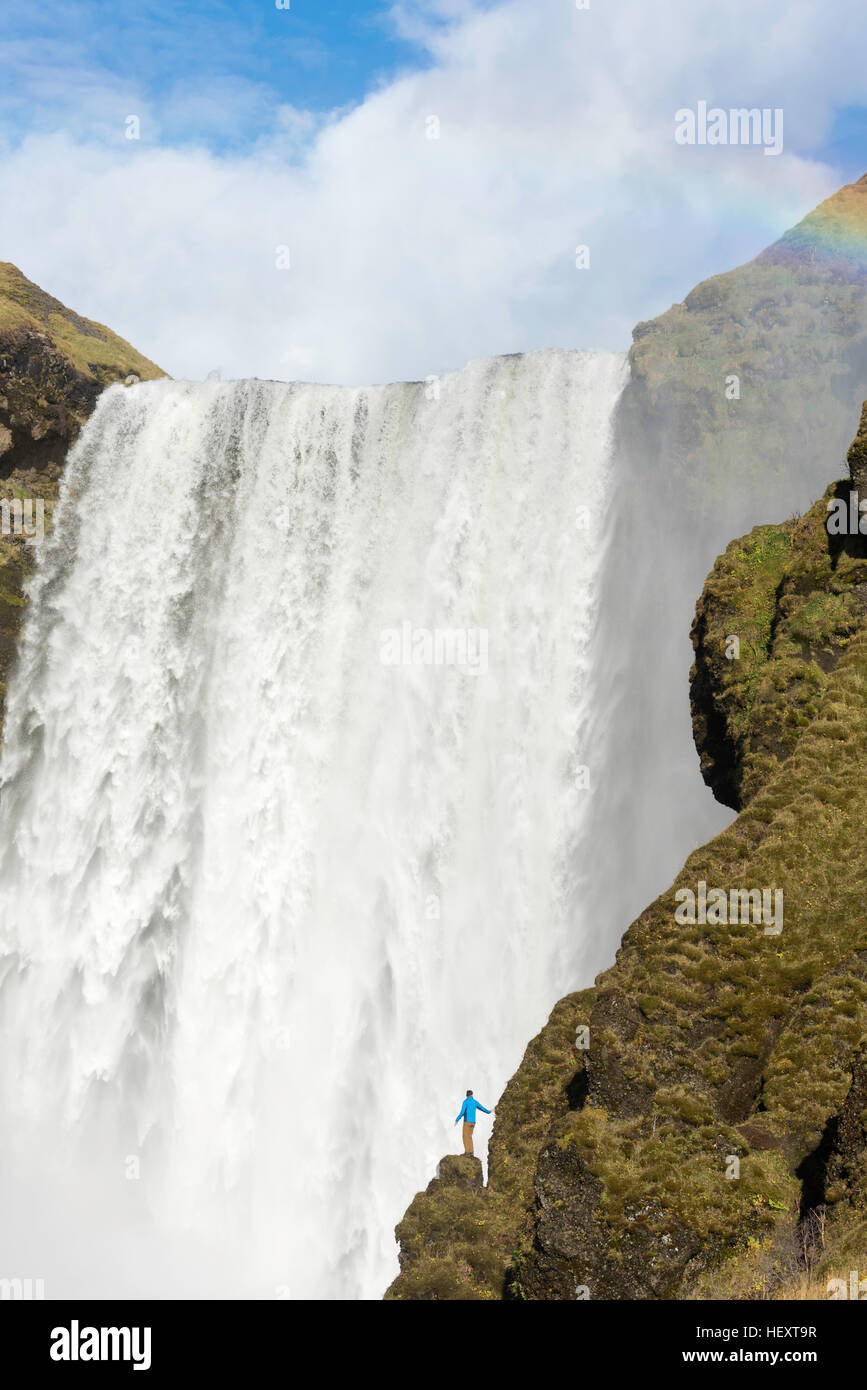 Giovane Uomo in camicia blu sorge su una scogliera con Skogafoss cascata dietro, Islanda. Foto Stock